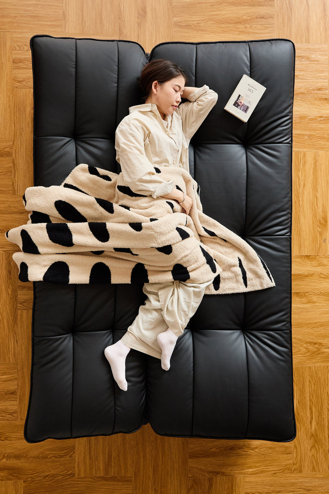 Person wrapped in a polka dot blanket on a black cushioned chair with wooden floor.