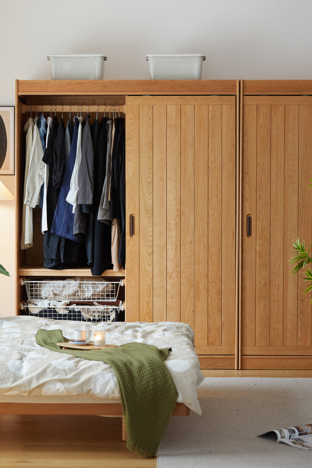 Wooden wardrobe with clothes inside, bed with green blanket, and light-colored walls.