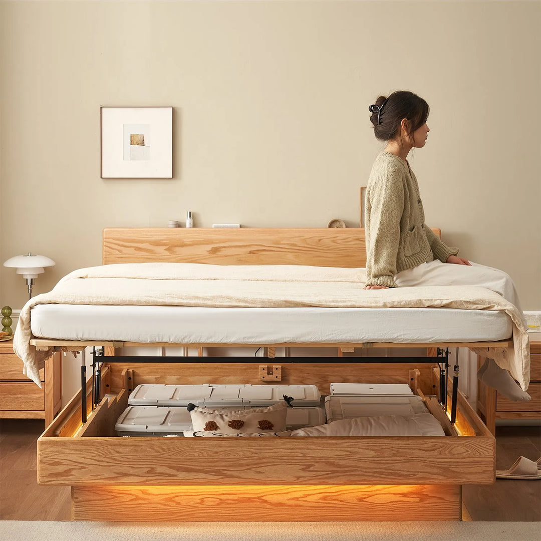 Loft Home oak wood bed frame with lift-up storage, featuring a woman sitting on the bed in a warm and cozy bedroom setting.