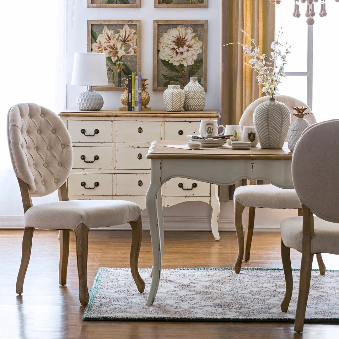 Rustic dining room setup featuring a distressed wooden table, upholstered chairs with tufted backs, and vintage decor elements including a dresser with floral artwork, perfect for a cozy and charming home interior.