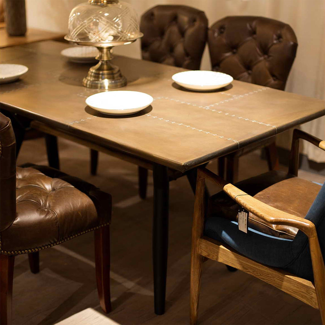 Loft Home brass dining table collection, showcasing a handcrafted brass tabletop with elegant stitching details and paired with luxurious leather chairs, creating a vintage-inspired dining atmosphere.