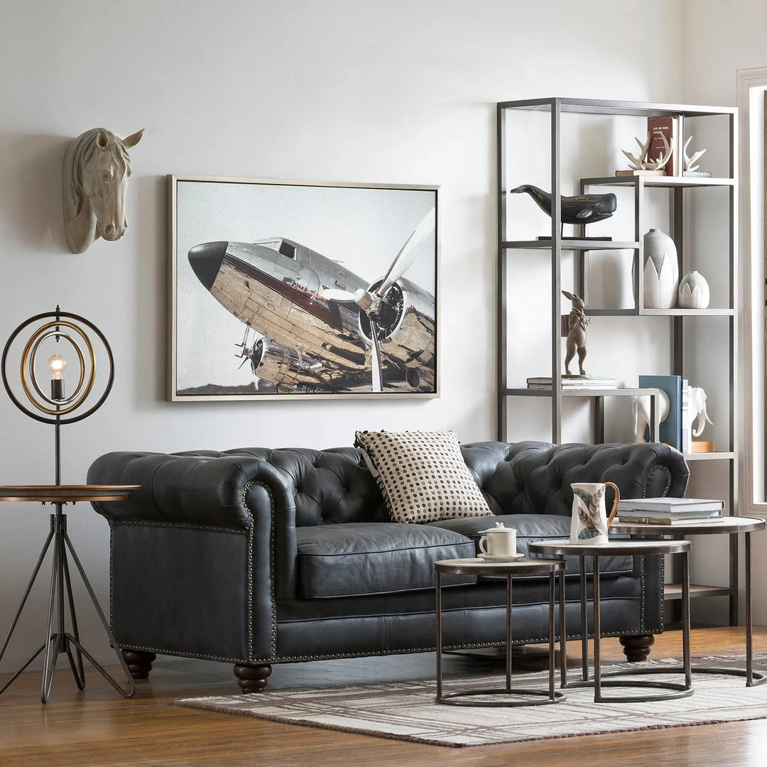 Industrial-style living room featuring a black leather chesterfield sofa, accent pillows, modern side tables, a unique lamp, and a wall shelf with decorative items. The decor includes a striking airplane artwork and a horse head sculpture.