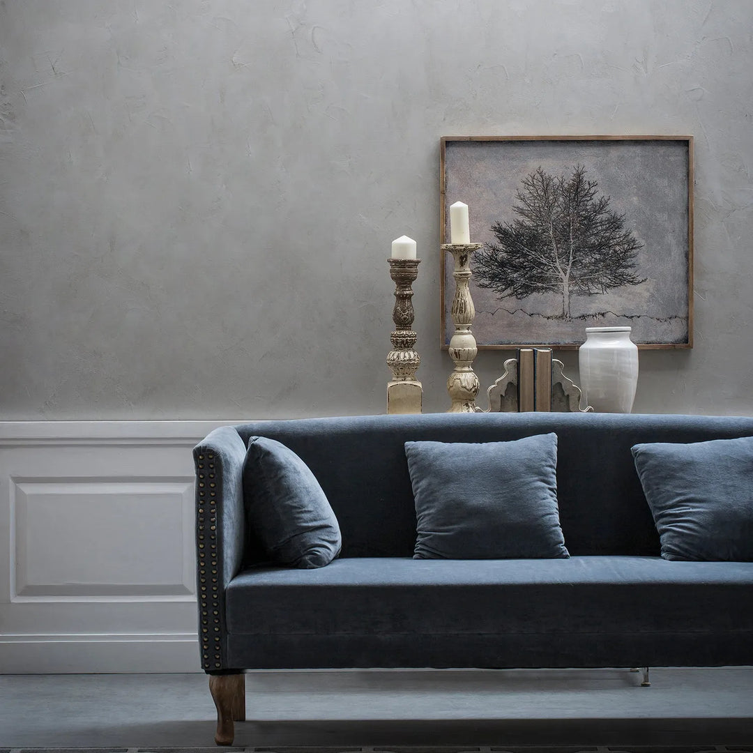 Elegant living room with a sleek grey sofa, adorned with matching cushions, set against a minimalist backdrop with decorative candlesticks, a ceramic vase, and a framed artwork of a tree.