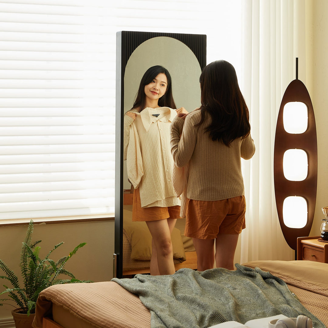 Bedroom scene with a woman trying on clothes in front of a full-length mirror, accompanied by soft lighting, natural decor, and cozy bedding.