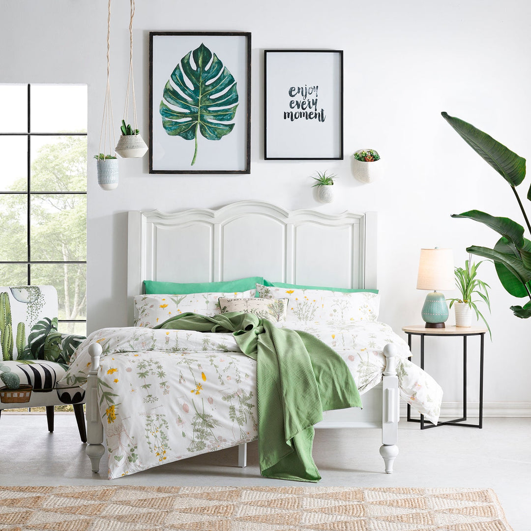 Bright bedroom with a white bed frame, botanical bedding, green accents, houseplants, and inspirational wall art, creating a fresh and serene ambiance.