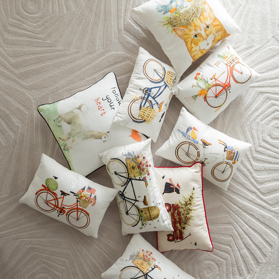 An assortment of decorative cushions, featuring various designs including bicycles with baskets of flowers, a cat, and a 'Follow your heart' message, displayed on a patterned rug.