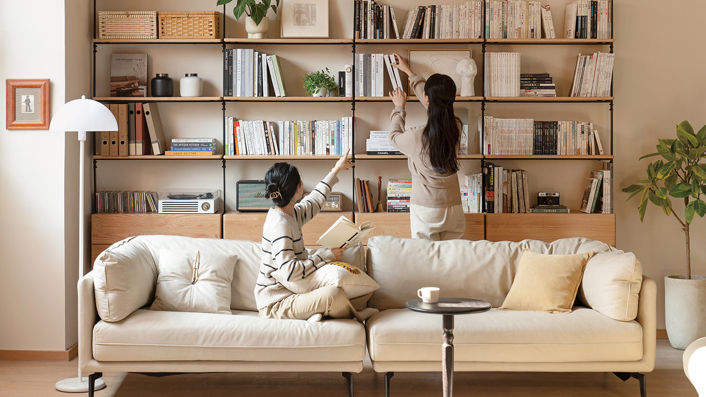 Two women organizing a bookshelf in a cozy living room with a white sofa and modern decor, perfect for relaxed and functional home spaces.