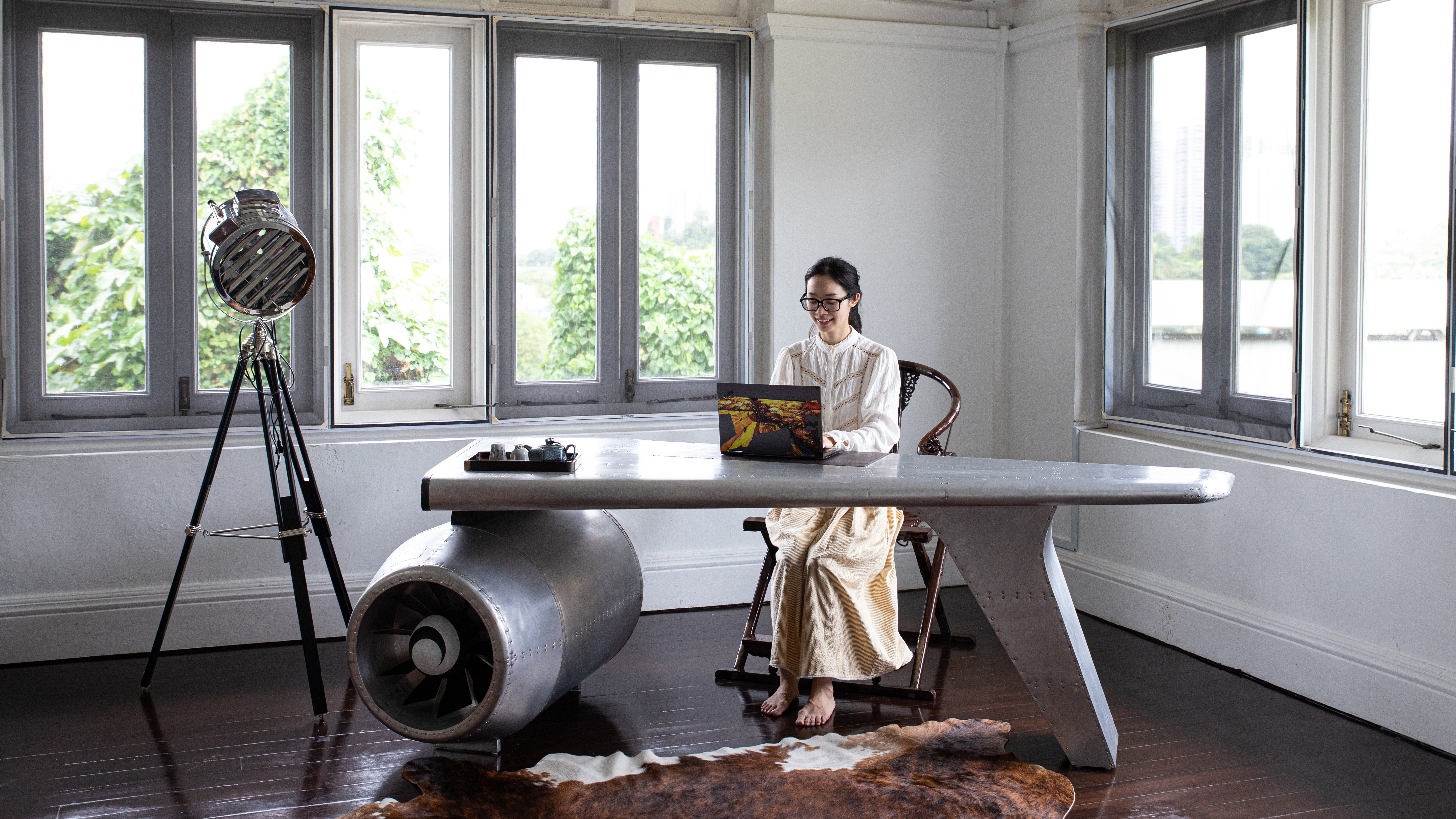 Loft Home industrial-style office featuring a woman working on a laptop at an airplane wing desk with large windows and natural light.