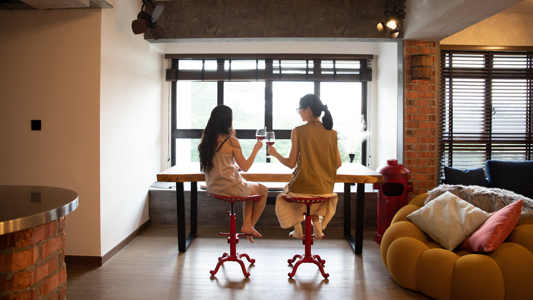 Loft Home stylish furniture featuring two women enjoying wine at a wooden table with red stools and a mustard sofa.