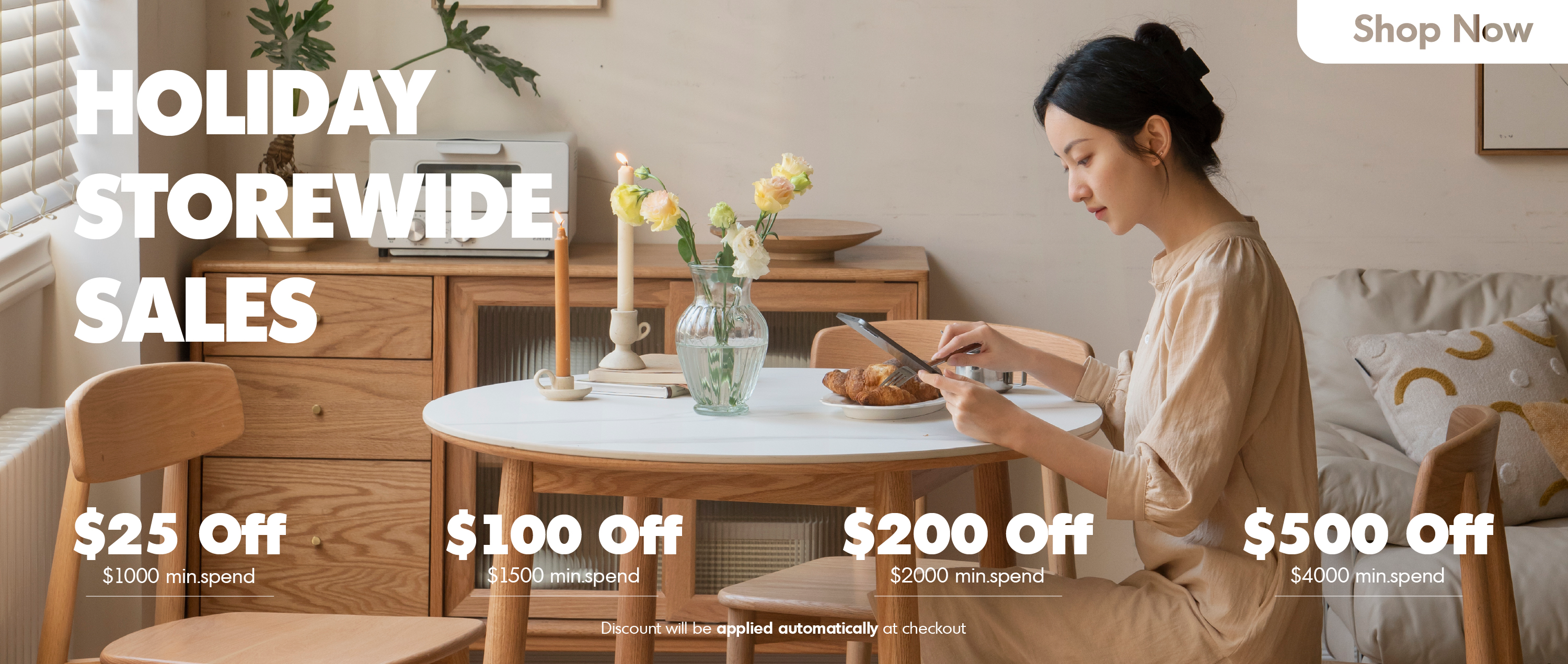 Elegant holiday dining setup with light wood furniture, woman browsing tablet, showcasing LOFT HOME storewide sales up to $500 off with minimum spend.