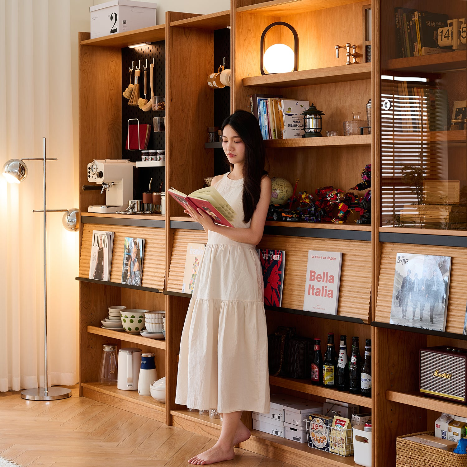 Cozy living space with wooden shelves, books, magazines, kitchen items, a floor lamp, and a woman in a white dress reading a book, creating a warm ambiance.